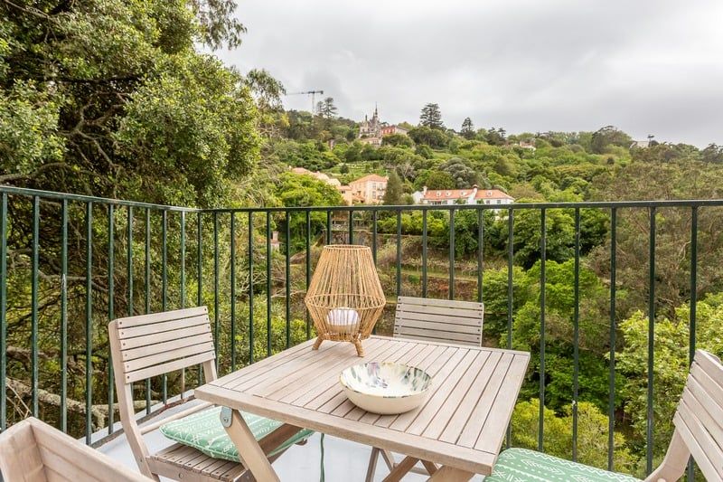 A slatted table on the terrace with a wicker lantern and a bowl on it, chairs drawn up, the valley behind.