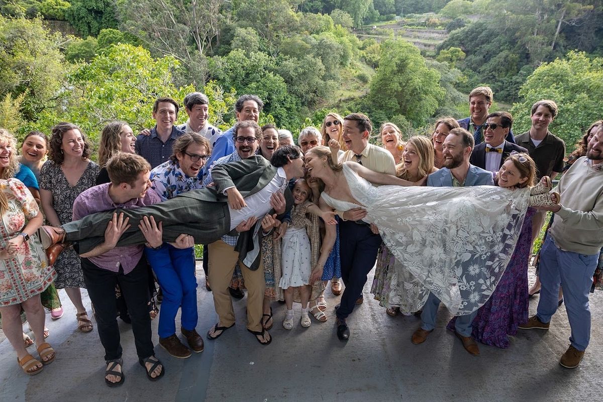 A wedding party crowded together in the garden, lifting the bride in her veil, everyone laughing.