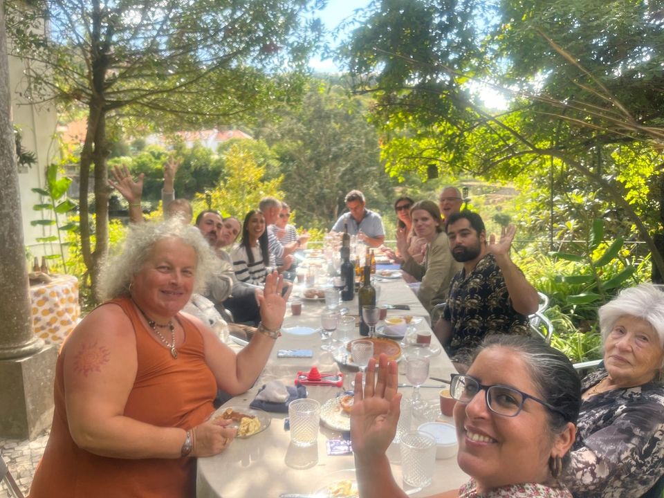 A party of guests waving at the camera from a long table set among the garden greenery.