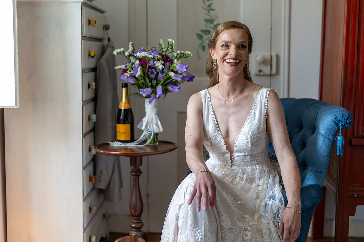 A bride in her lace dress sitting and laughing in one of the bedrooms, her bouquet and a bottle of champagne on the table beside her. Photograph by Chas Studios.