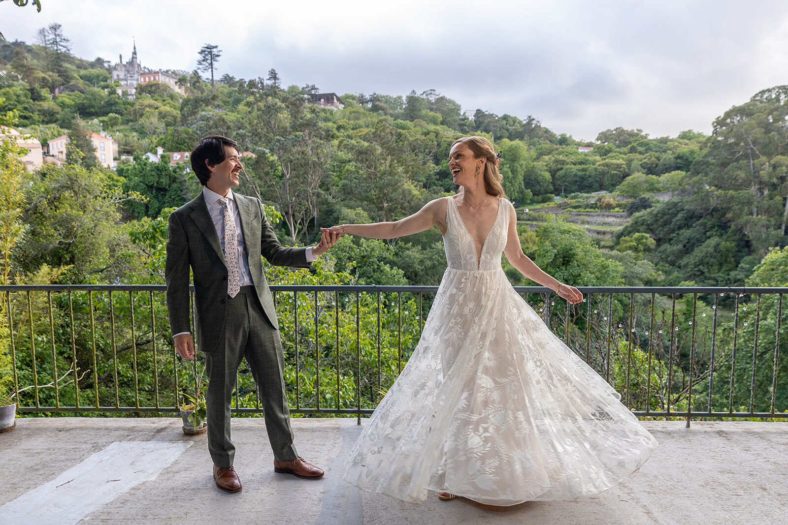A bride and groom on the terrace at Casa da Paderna, her skirt turning as she steps away from him, both of them laughing, with the wooded Arra&ccedil;&aacute;rio Valley and the hillside of Sintra behind them.