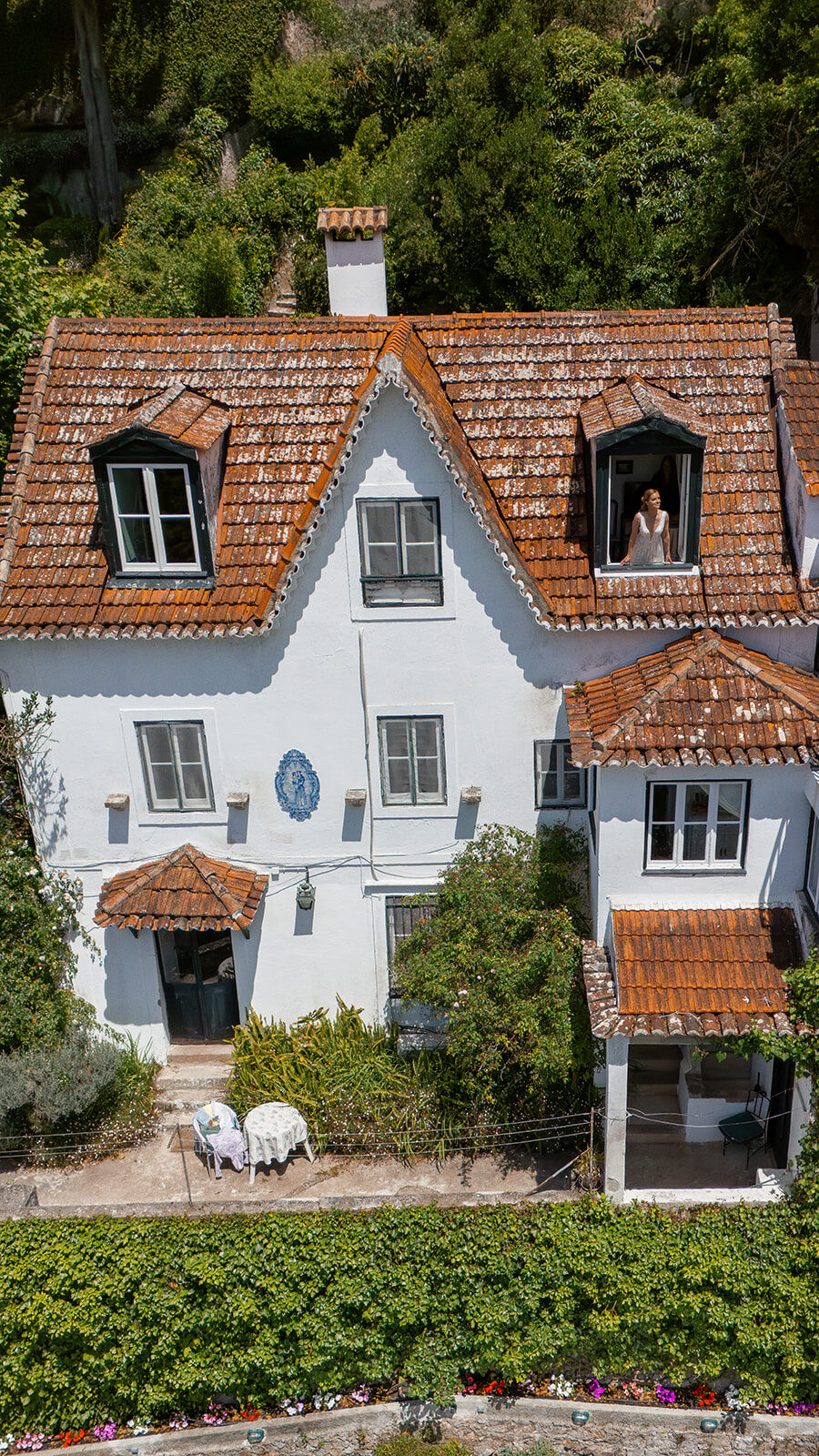 Casa da Paderna seen head-on from the air: whitewashed walls, scalloped eaves and terracotta roofs banked into the hillside, with the bride leaning out of an attic window in her dress.