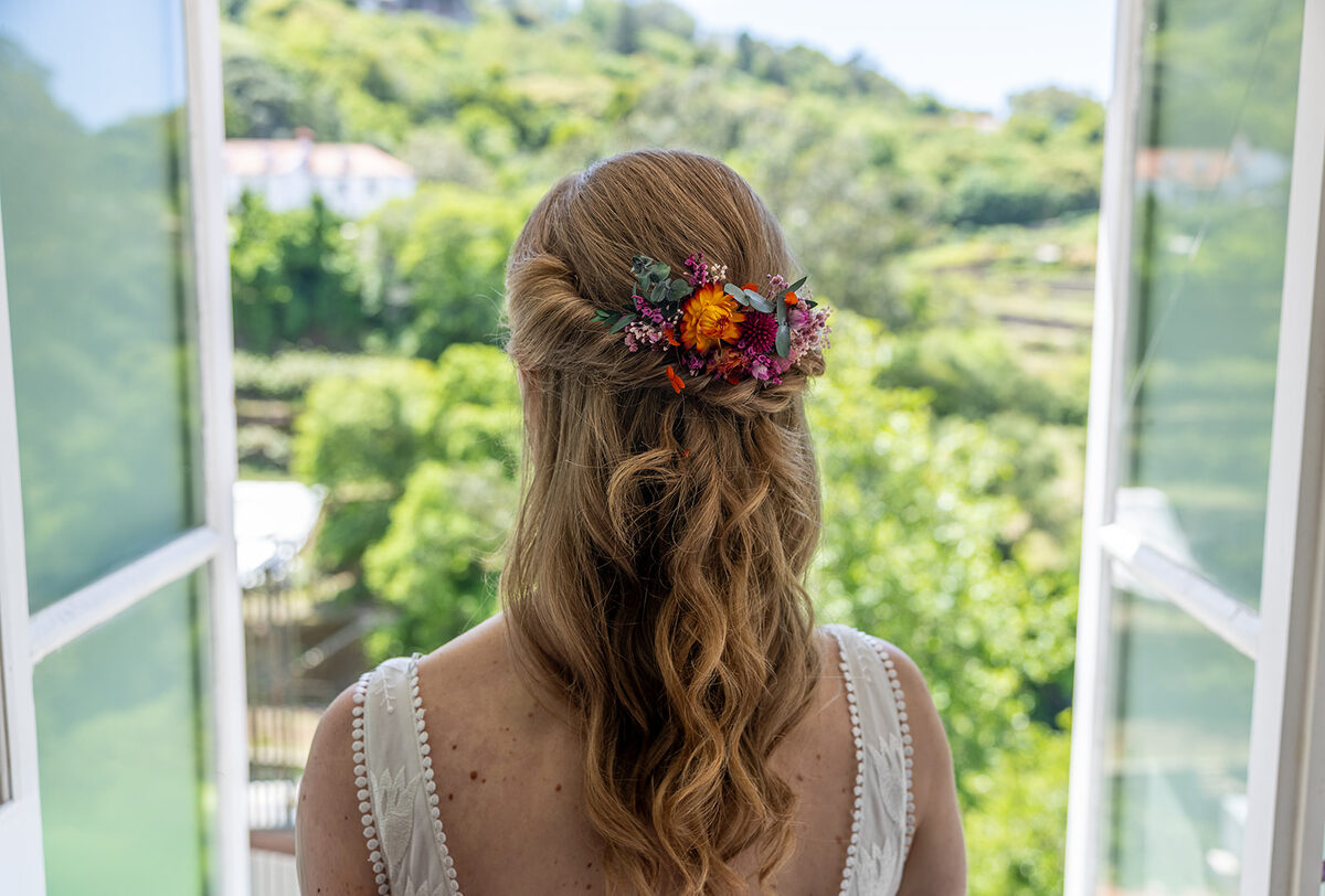 A bride seen from behind at an open window in the house, dried flowers pinned into her hair, looking out over the terraced Arra&ccedil;&aacute;rio Valley.