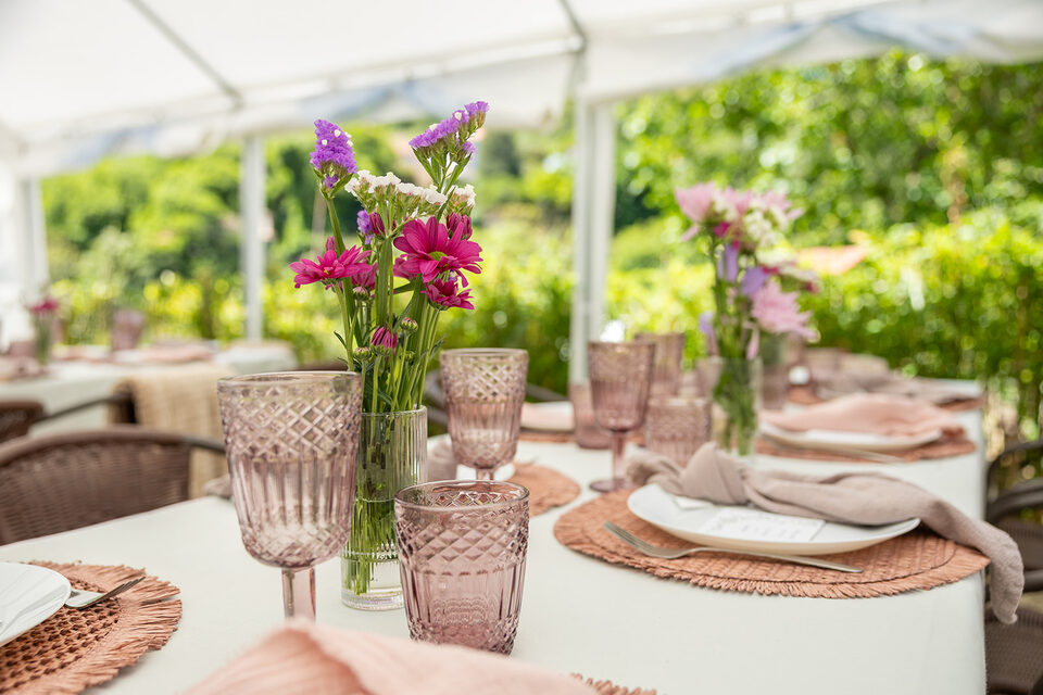 A wedding table laid under the marquee in the garden: rose-coloured cut glass, linen napkins on rattan placemats and jars of garden flowers, with the green of the valley beyond the open sides.