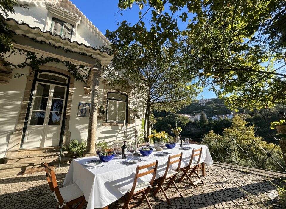 A long table laid for dinner on the terrace outside the front door of Casa da Paderna, white cloth and blue-and-white plates, with the roofs of Sintra Old Town on the hillside beyond.