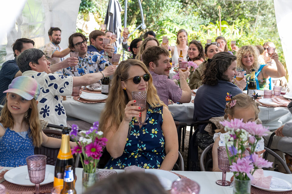 Wedding guests along the long table under the marquee, grandparents and small children among them, raising rose-coloured glasses to a toast.