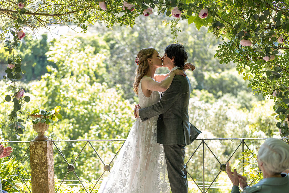 A bride and groom kissing under an arch of roses and eucalyptus in the garden at Casa da Paderna, the wooded valley behind them and guests applauding at the edge of the frame.